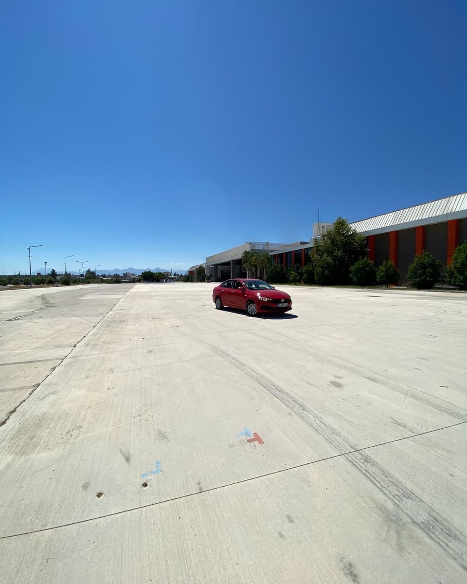 Aksu'daki geniş beton alanda park etmiş kırmızı Fiat Egea; arkada Toros Dağları ve açık mavi gökyüzü