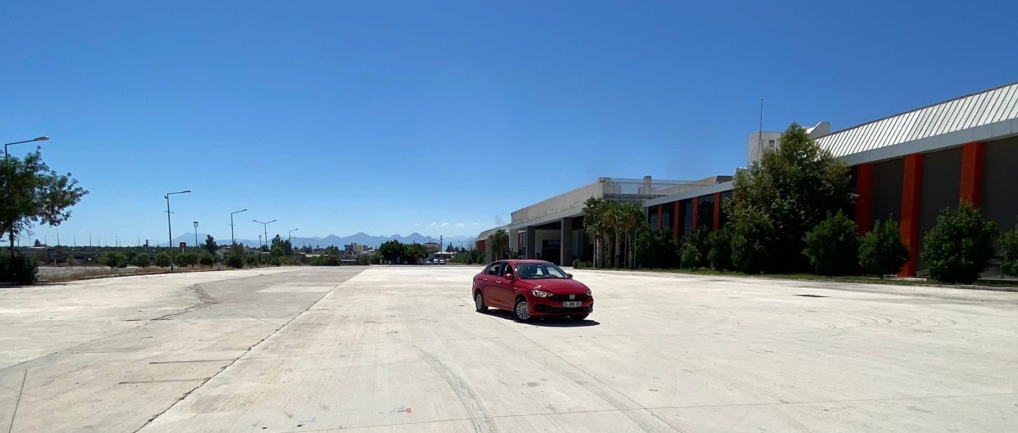 Aksu'daki geniş beton alanda park etmiş kırmızı Fiat Egea; arkada Toros Dağları ve açık mavi gökyüzü